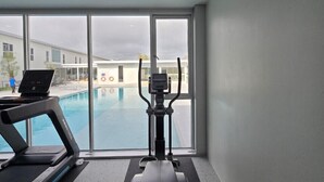 Une salle de sport équipée d’un tapis de course et d’un vélo elliptique, avec de grandes fenêtres donnant sur une piscine.