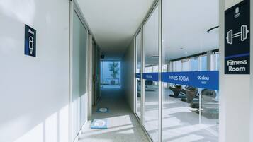 Pasillo que conduce a salas de gimnasia con señalización clara, una puerta de cristal y vistas a equipamiento de ejercicio exterior.
