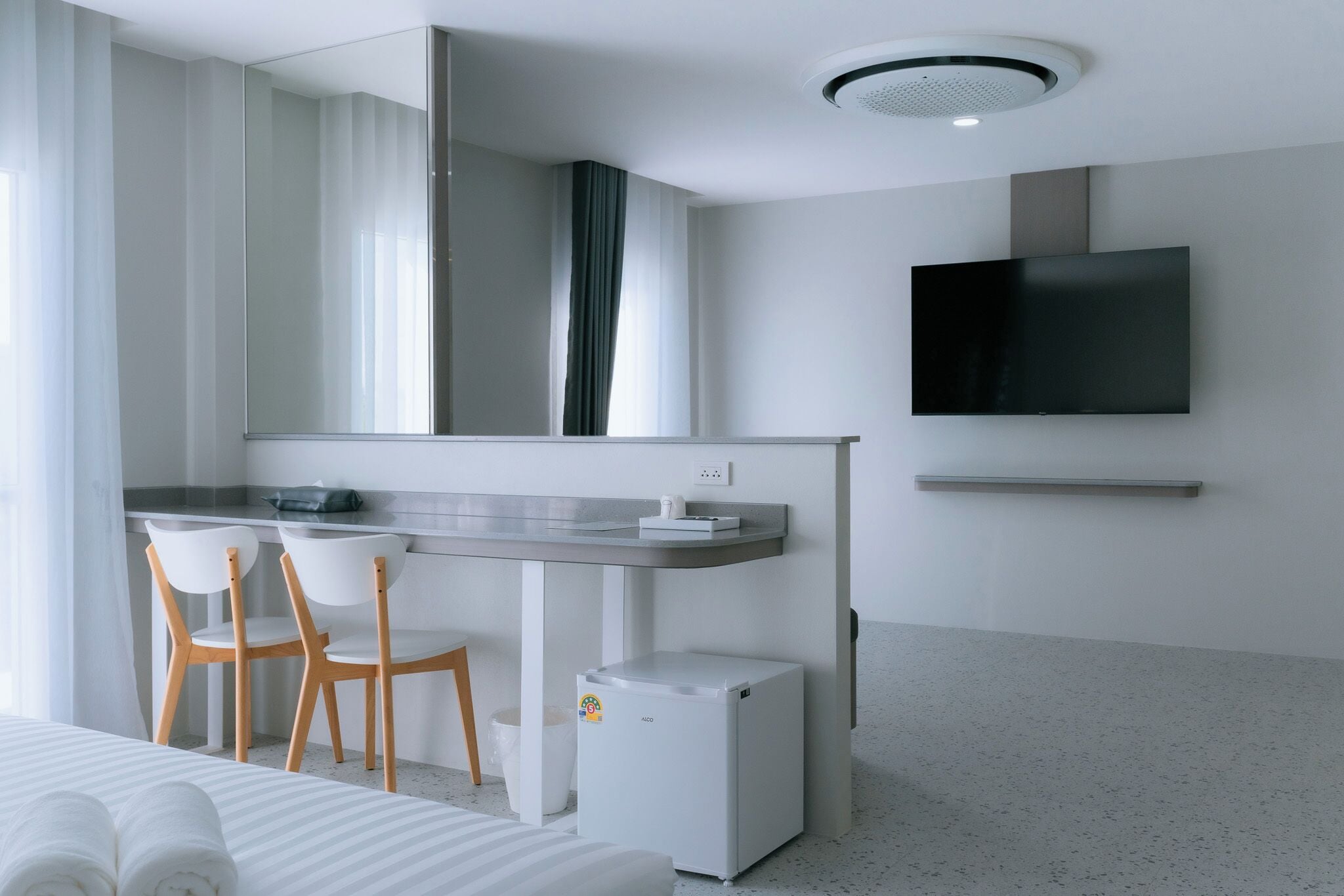Una habitación de hotel moderna con una cama, un escritorio con dos sillas, un televisor en la pared y un refrigerador pequeño.