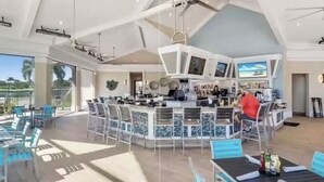 A spacious bar area with a high ceiling and a large TV screen. There are multiple seating areas with tables and chairs, and a pool area is visible in the background.
