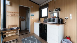 Una cocina compacta con microondas, cafetera y un lavamanos pequeño. Hay una mesa de madera con un bol y una silla. Se observa un extintor en la esquina.
