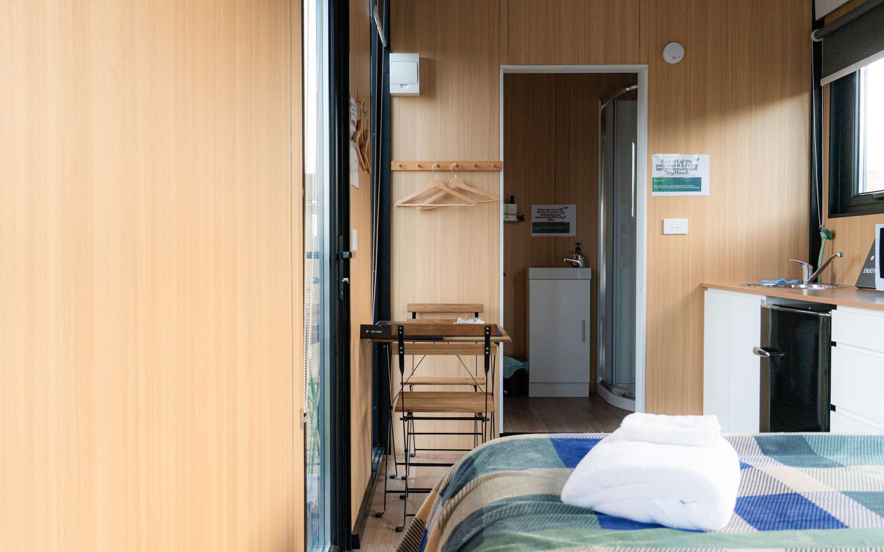 Une petite pièce comprenant un lit, une petite table, une chaise, un porte-manteau mural, un lavabo et une kitchenette compacte.
