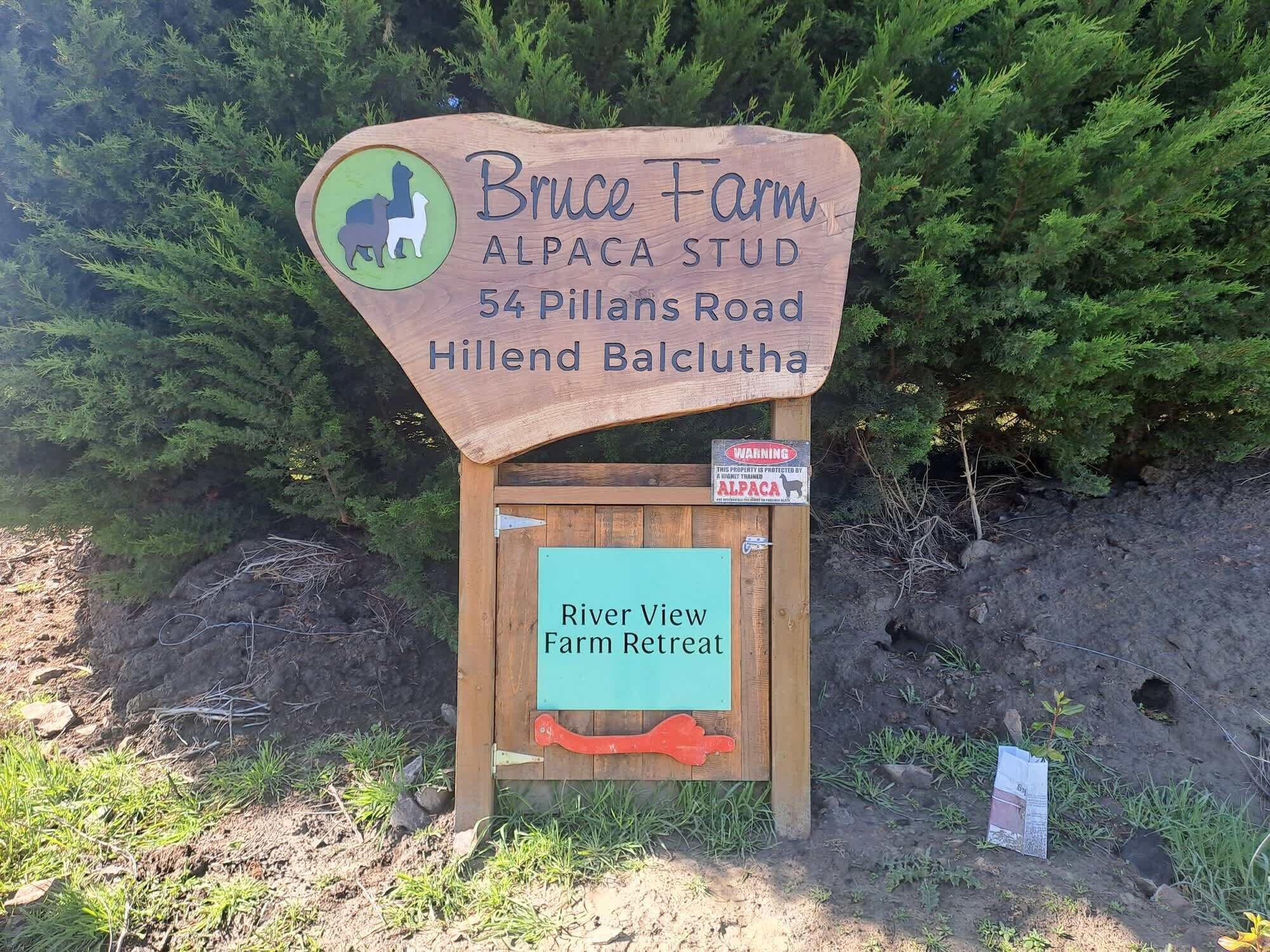 Ein Holzschild für Bruce Farm Alpaca Stud in der Pillans Road 54, Hillend Balclutha, das einen Flussblick-Bauernhofurlaub anwirbt.