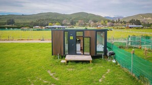 Une cabane moderne et compacte, dotée d’une façade en bois et de portes en verre, située dans un champ verdoyant, offrant une vue sur des collines ondulantes et, au loin, sur un terrain de sport clôturé.