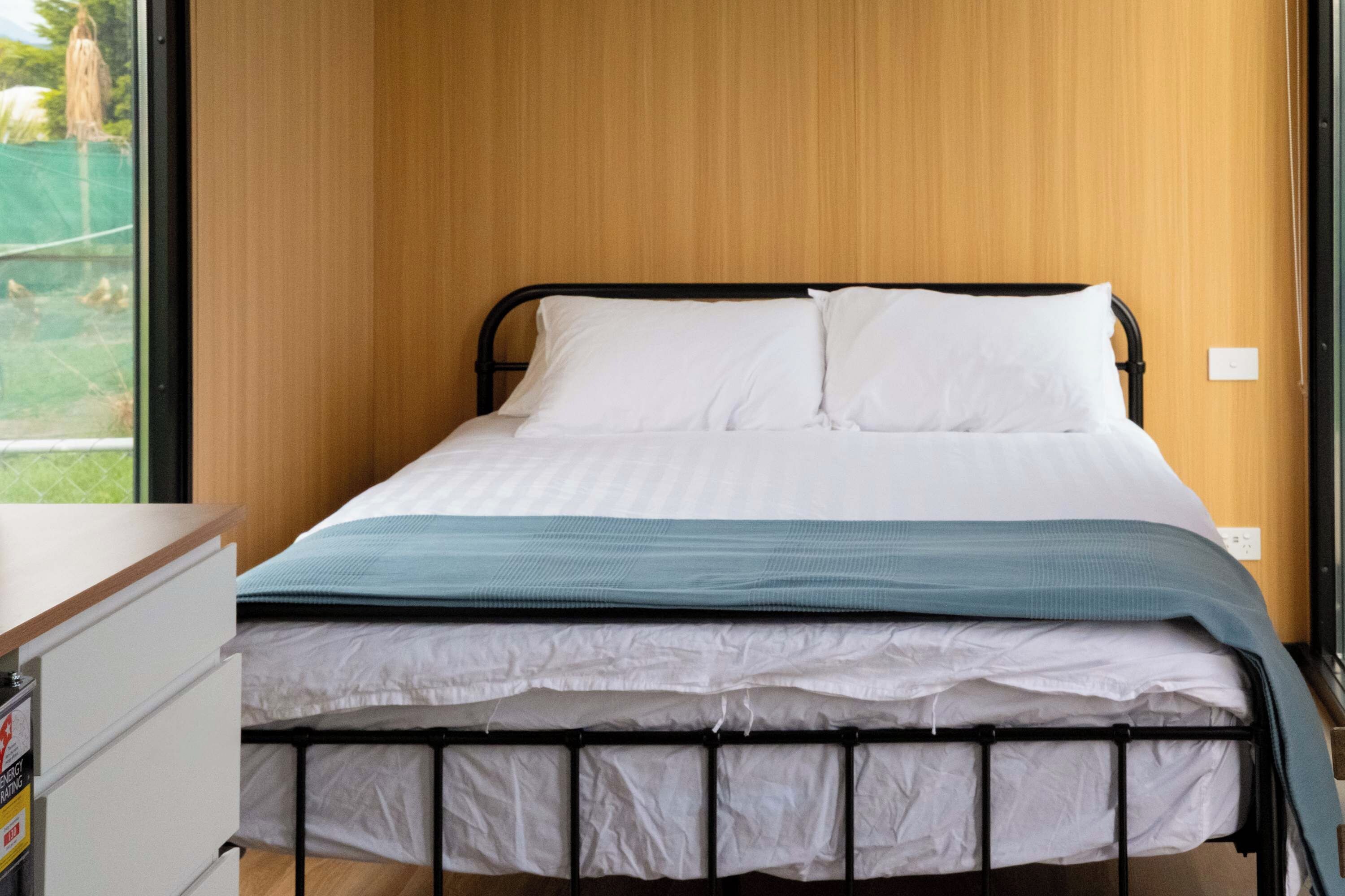 Un lit bien fait, avec une literie blanche et bleue, un mur en bois derrière et une fenêtre sur la gauche.