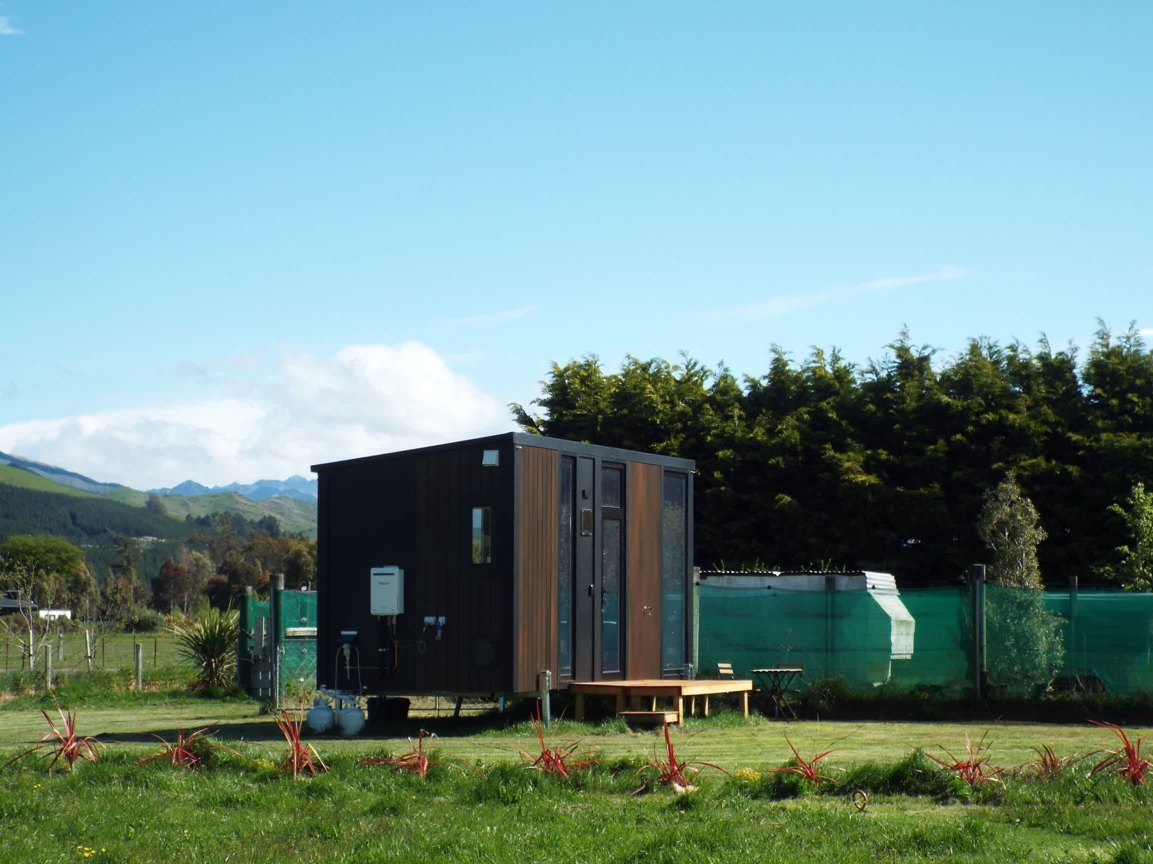 A modern, compact cabin with a flat roof, situated in a grassy field with a view of distant mountains.