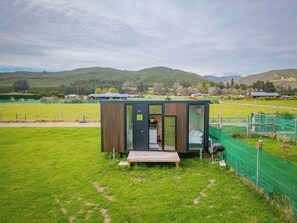 Une cabane moderne et compacte, dotée d’une façade en bois et de portes en verre, située dans un champ verdoyant, offrant une vue sur des collines ondulantes et, au loin, sur un terrain de sport clôturé.