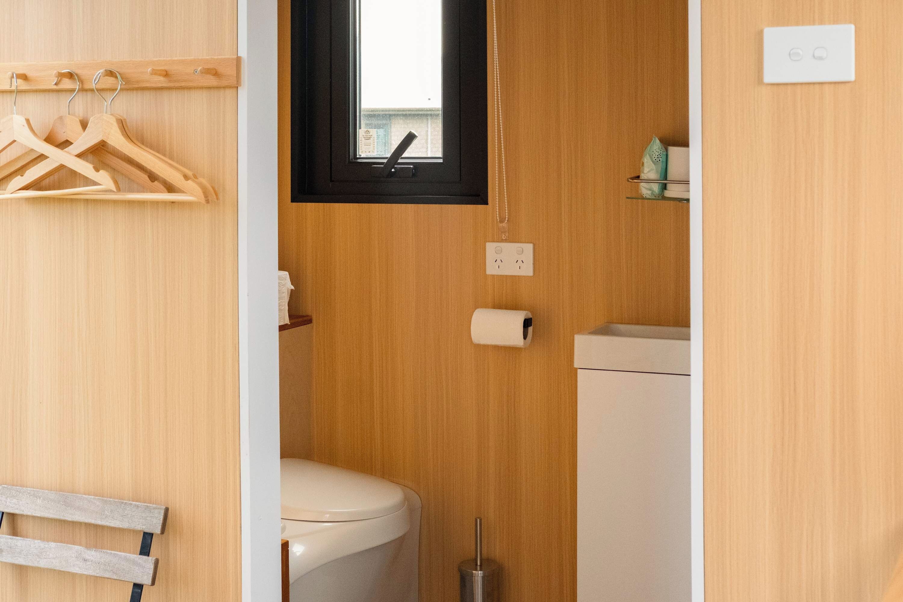 A compact bathroom with a toilet, a small window, a wooden wall, and a shelf with toiletries.