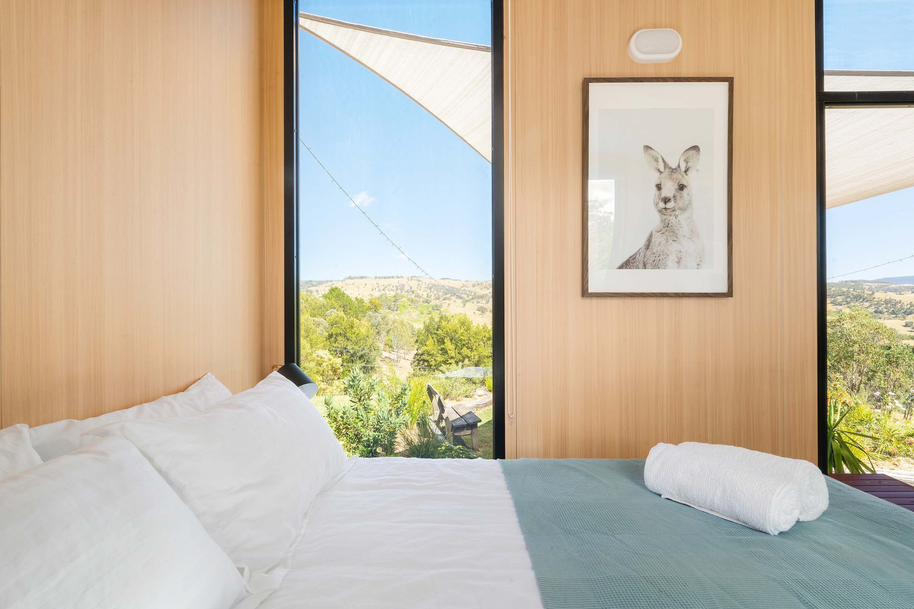 Een slaapkamer met een bed, een prent van een kangoeroe en uitzicht op groen door grote ramen.