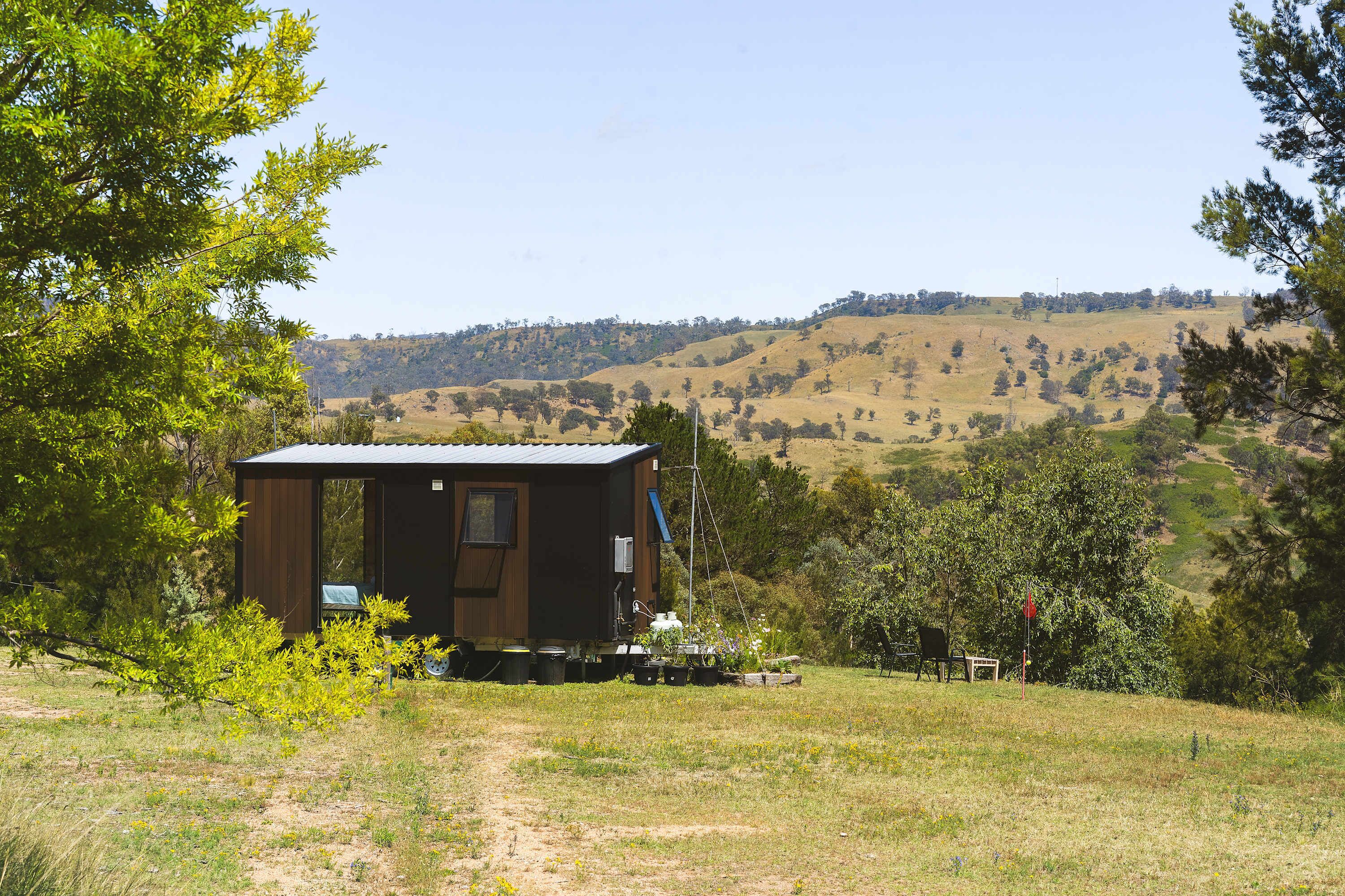 A small cabin with a metal roof, surrounded by a grassy field and trees, with a hilly landscape in the background.