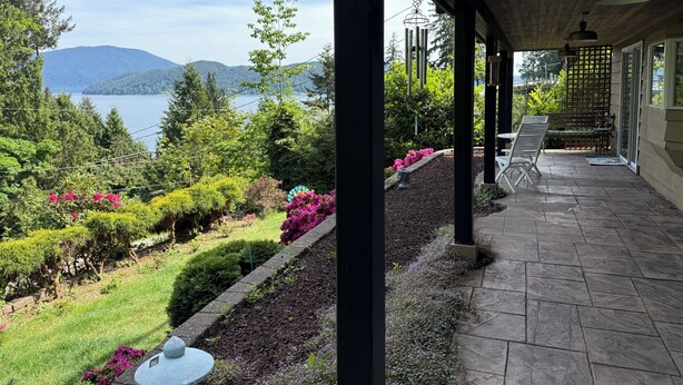 A covered outdoor area with a view of a lake and mountains, stone flooring, and a seating area with chairs and a table.