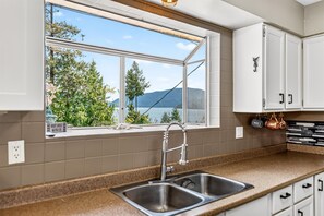 A kitchen with a large window, a stainless steel sink, and white cabinets. - Cozy Home with Ocean and Mountain views. Family sized wth game room and deck (Gibsons)