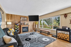 Un salon avec une cheminée, un canapé, une table basse et une grande fenêtre donnant sur les arbres.