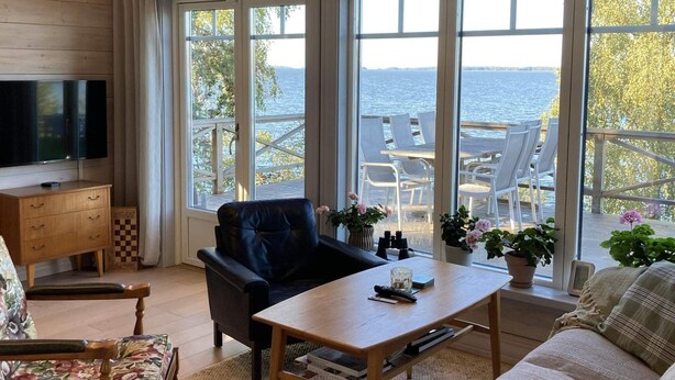 Un salon confortable avec un téléviseur, un fauteuil en cuir, une table basse en bois et un canapé. De grandes fenêtres offrent une vue sur l’eau et sur les chaises longues situées à l’extérieur.