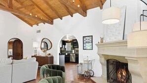A spacious living room with a fireplace, wooden ceiling beams, and a kitchen in the background. - Villa Sol: Private Las Palmas Estate Home (Palm Springs)