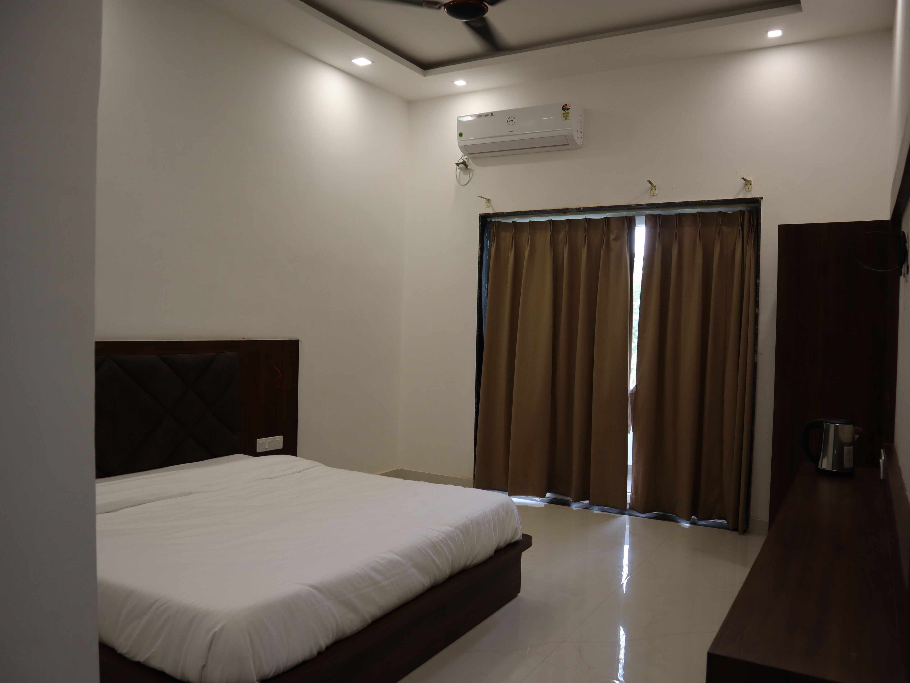 A bedroom with a bed, a ceiling fan, an air conditioning unit, a window with curtains, and a wooden nightstand.