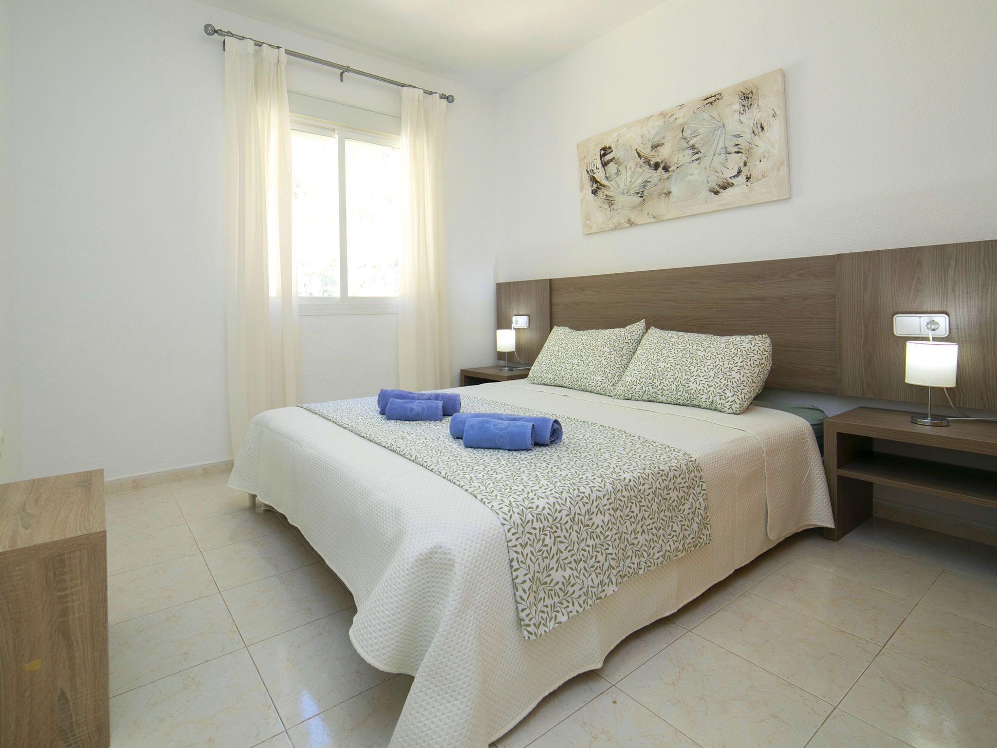 A hotel room with a large bed, two bedside tables with lamps, a window with curtains, and a wall-mounted abstract painting.