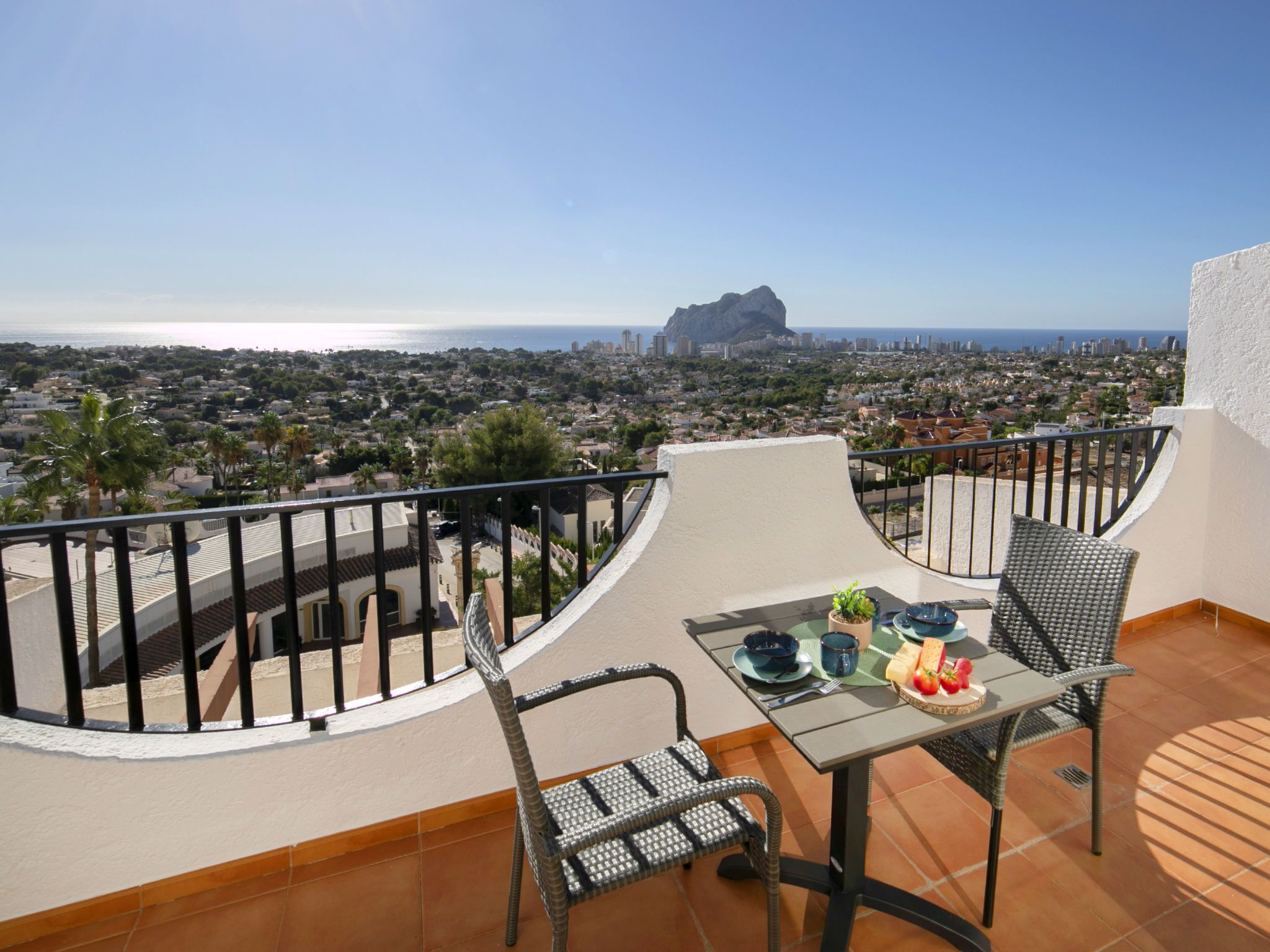 Un balcon avec une table dressée pour deux personnes, offrant une vue sur le paysage urbain côtier et sur une montagne imposante qui se profile à l’horizon.