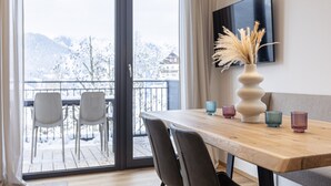Un espace repas moderne avec une table en bois, des chaises et un vase contenant des plantes séchées. Une grande fenêtre offre une vue sur un paysage enneigé.