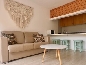 Uma sala de estar com um sofá bege, almofadas florais, uma mesa redonda e um painel de macramé na parede.