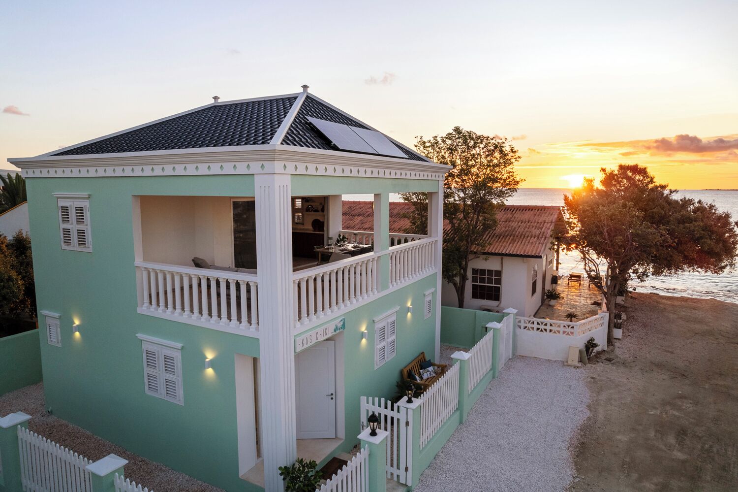 A two-story building with a balcony, a small attached structure, and a view of the sunset over the water.