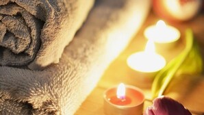 A wooden table with a rolled gray towel, two lit candles, and a purple flower. - Costa de Leticia (Alegria)