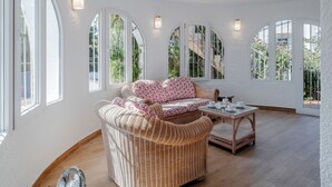 Ein Zimmer mit einem Korbssofa, einem Glastisch und bogenförmigen Fenstern mit Blick auf Grünflächen.