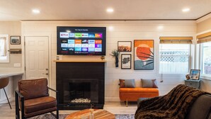 Una sala de estar con chimenea, un televisor de pantalla plana mostrando varios servicios de streaming, un sillón de cuero marrón, una mesa de centro de madera y un sofá gris con un plaid de piel.