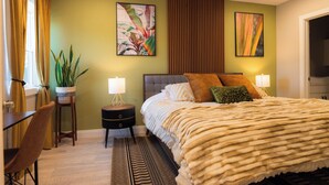 Un dormitorio con una cama grande, dos lámparas de noche, una planta en maceta, una mesita de noche de madera, una silla y un tapete estampado.