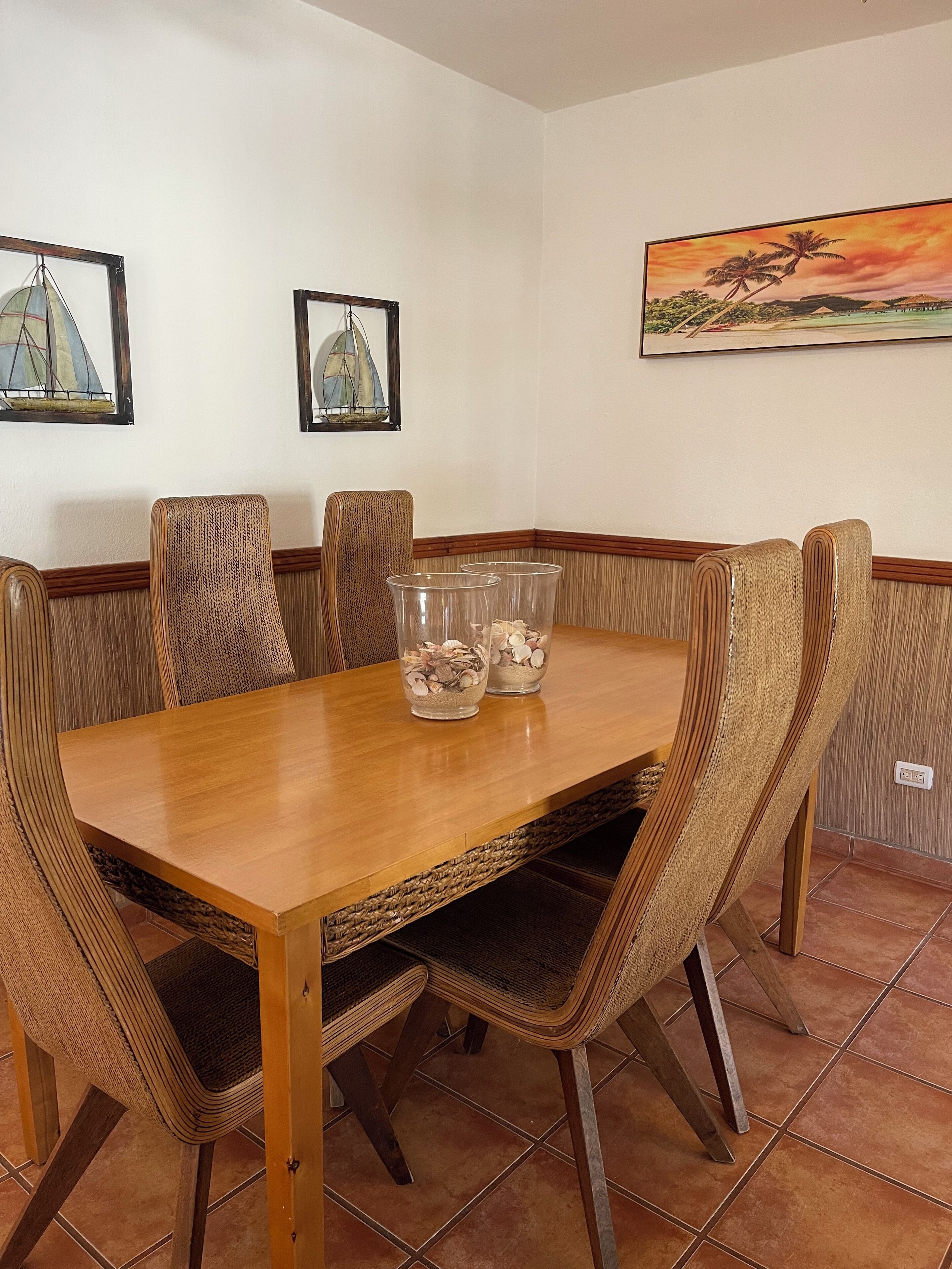 Uma área de refeições com mesa de madeira e cadeiras de fibra, dois vasos de vidro transparente com conchas e obras de arte emolduradas nas paredes.
