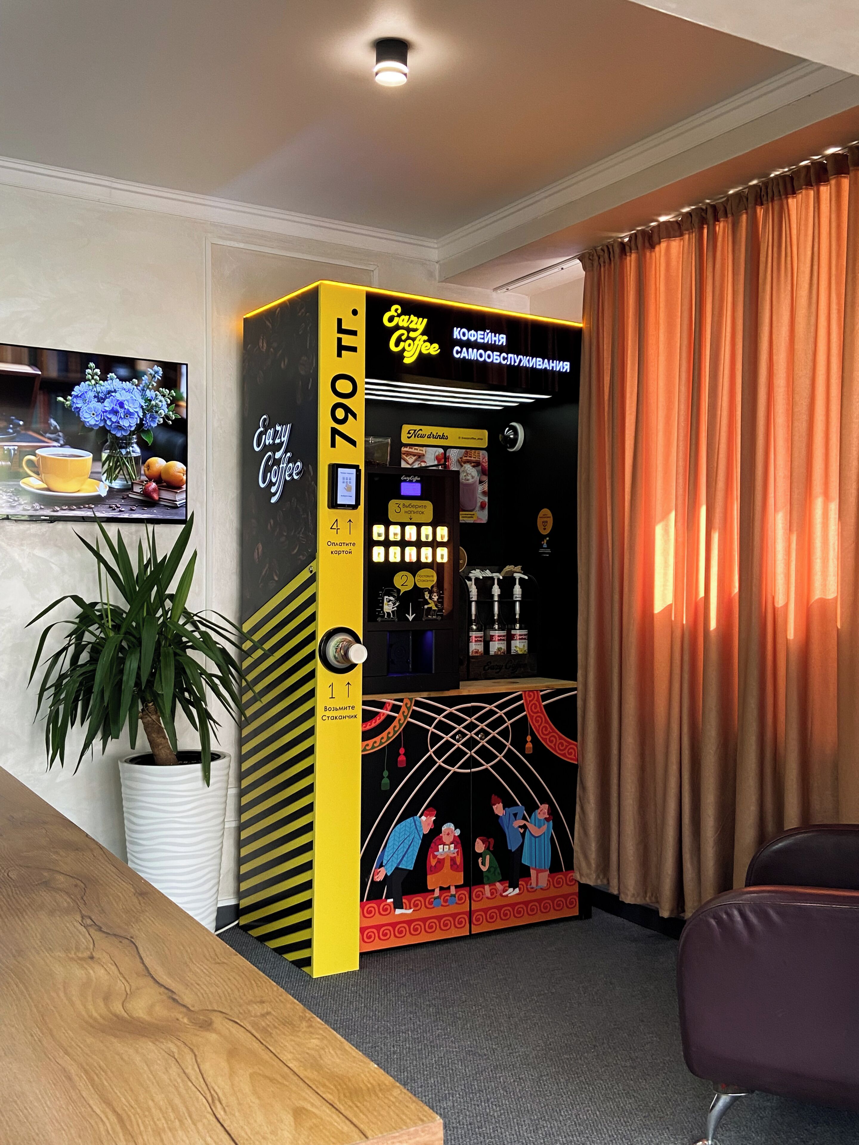A coffee vending machine with a digital interface, a potted plant, and a television on the wall.