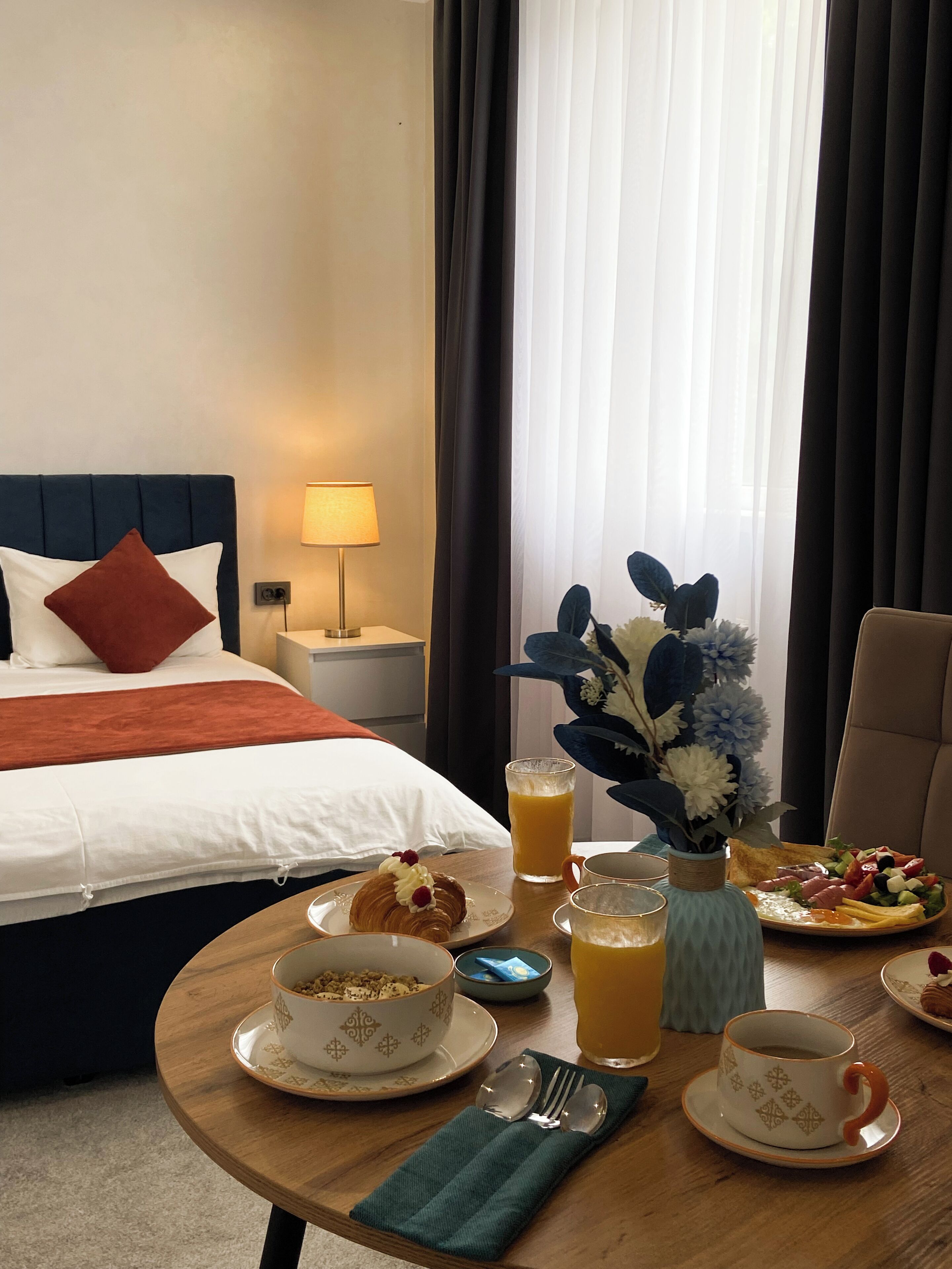A hotel room with a neatly made bed, breakfast table with plates of food, a vase with flowers, and a lamp.
