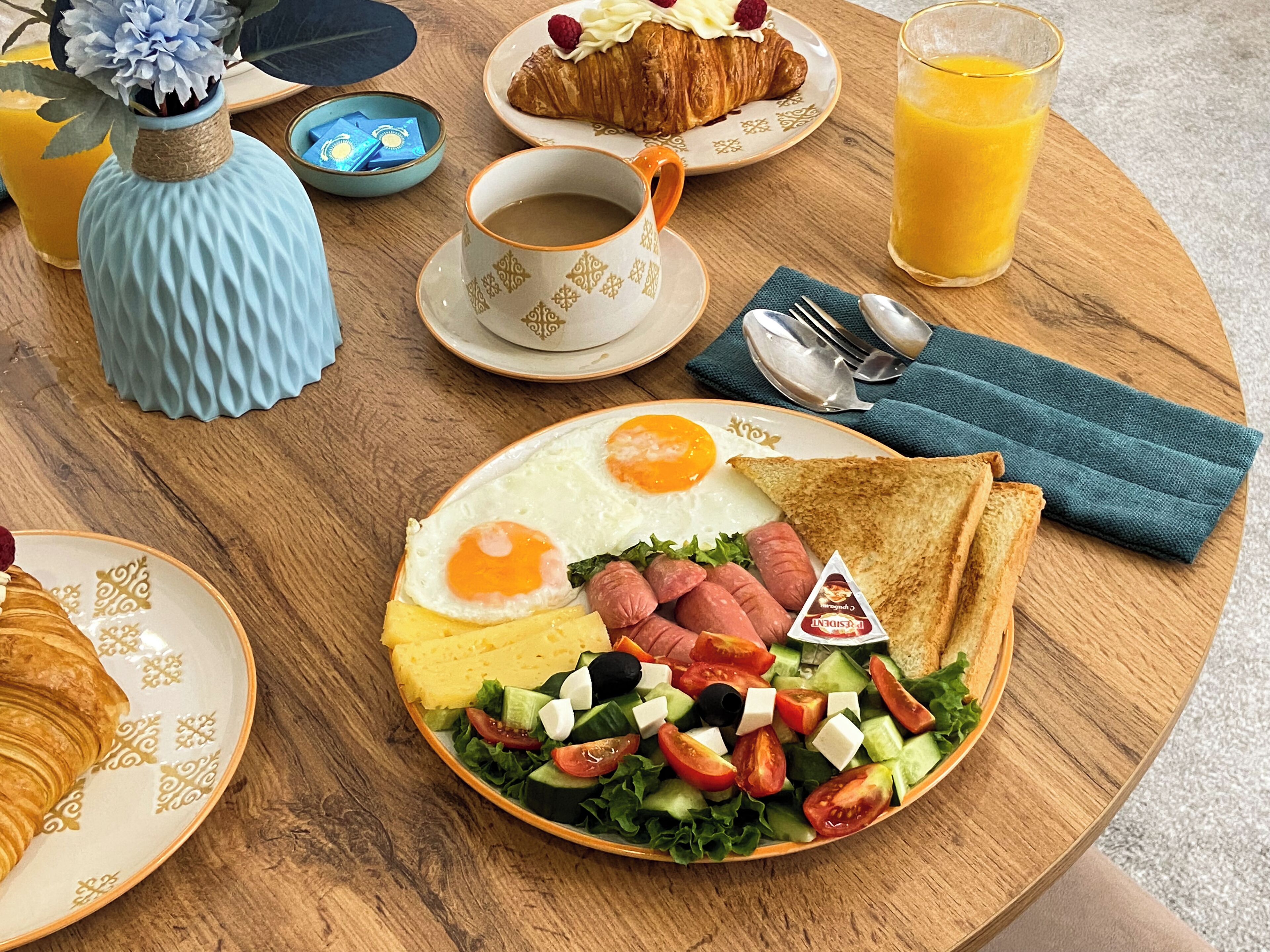 Une table en bois dressée pour le petit-déjeuner, avec des croissants, des œufs, des saucisses, une salade et un verre de jus d’orange.