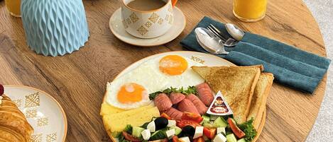 Desayuno con cruasanes, huevos, salchichas, una ensalada y un vaso de jugo de naranja sobre una mesa de madera.