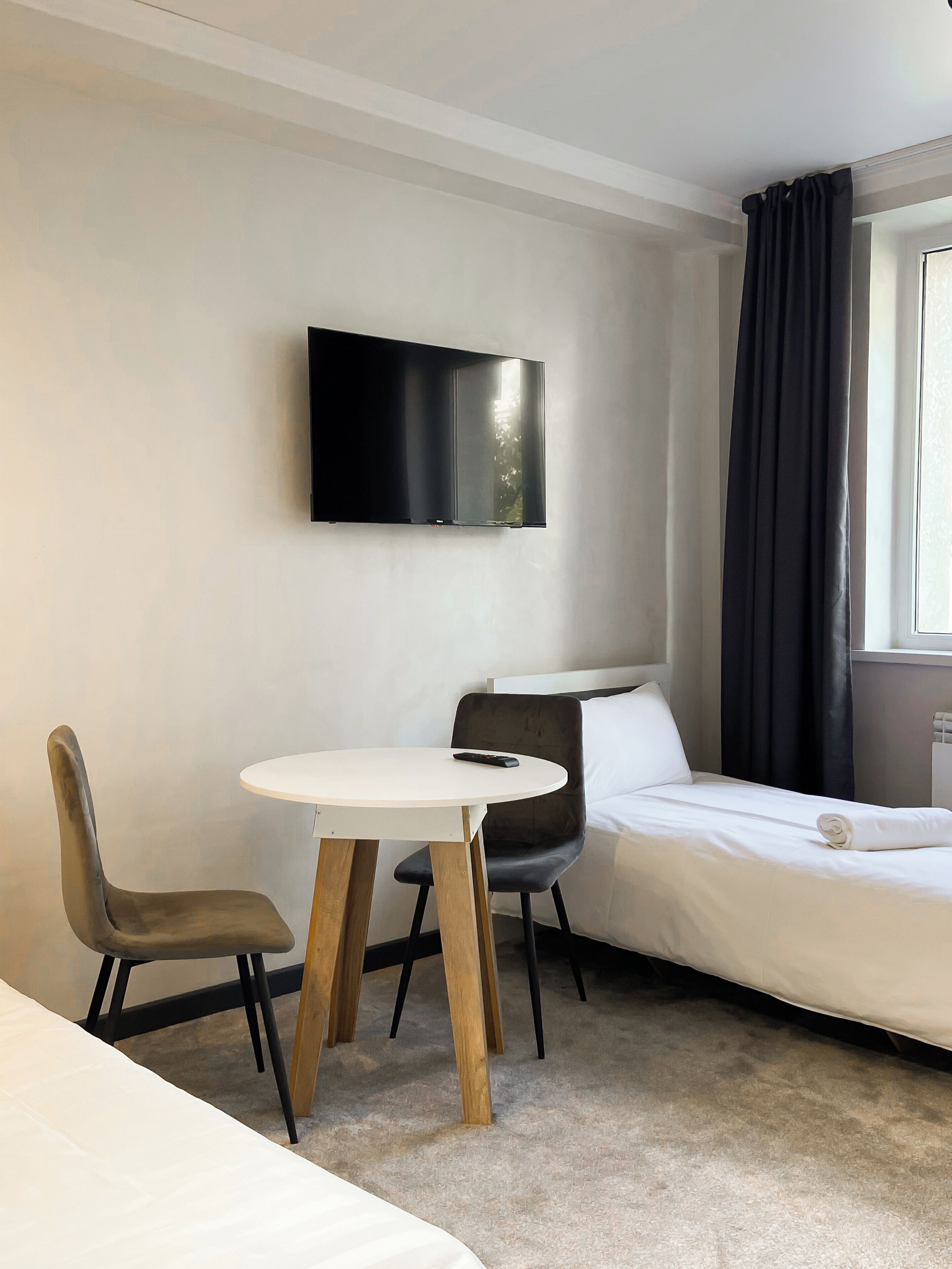 A modern hotel room with a bed, a TV mounted on the wall, a round table with two chairs, and a window with curtains.