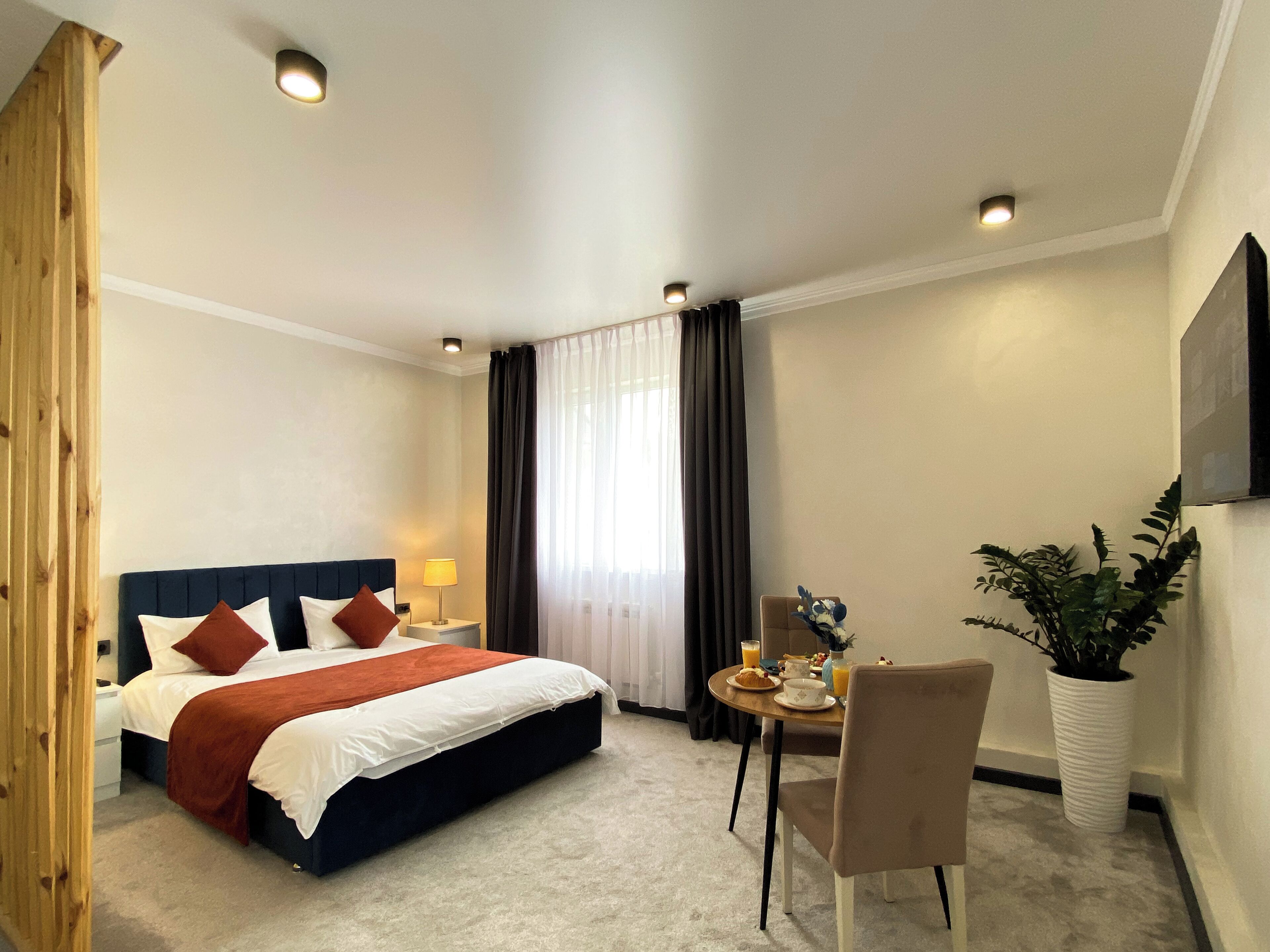 Habitación de hotel con cama, mesita de noche, lámpara, cortinas, una mesa de comedor con sillas, una planta en maceta y un televisor montado en la pared.