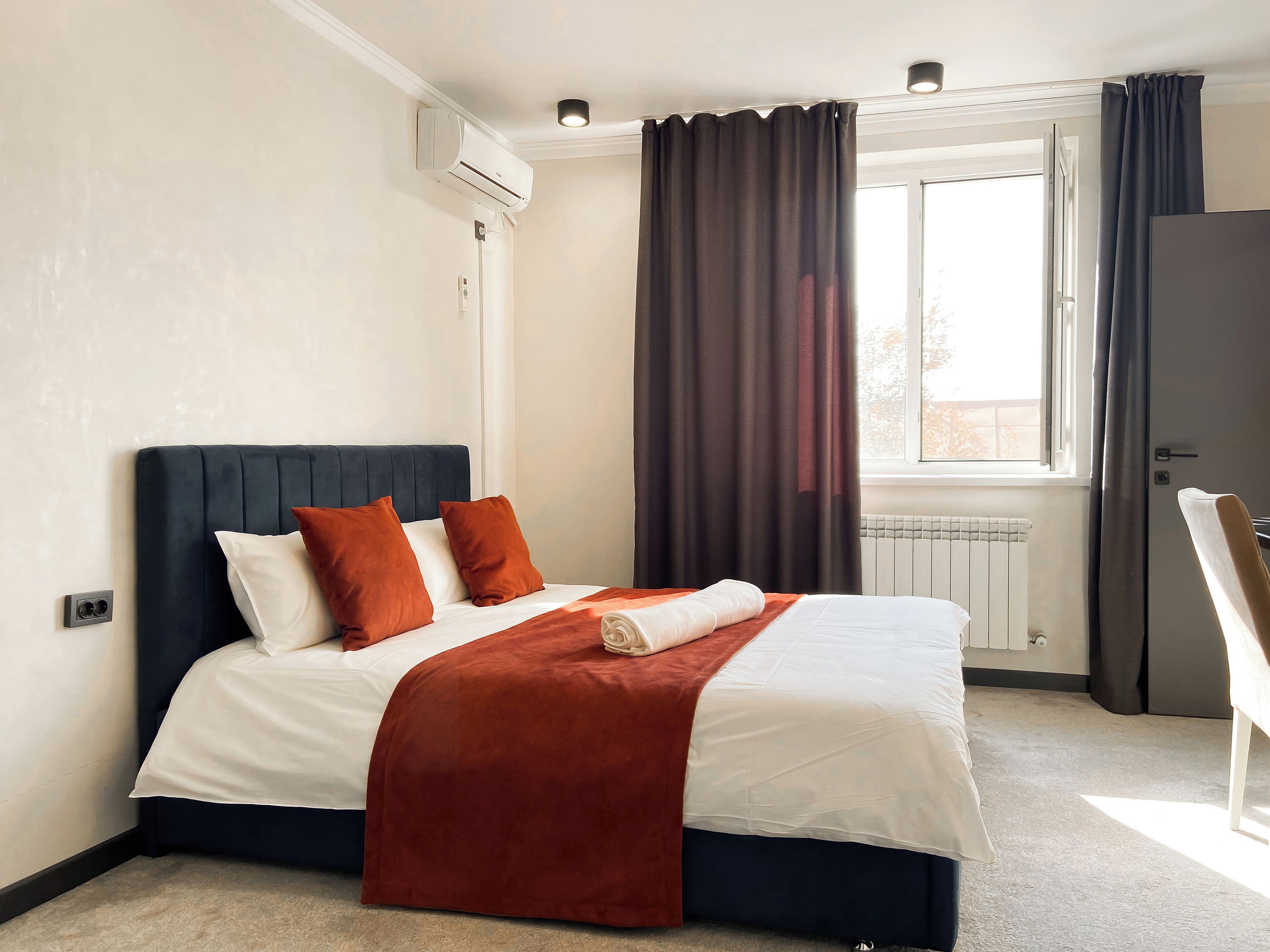 A neatly made bed with a dark headboard, white and orange pillows, and a red throw blanket. There is a window with dark curtains, a radiator below, and a door to the right.