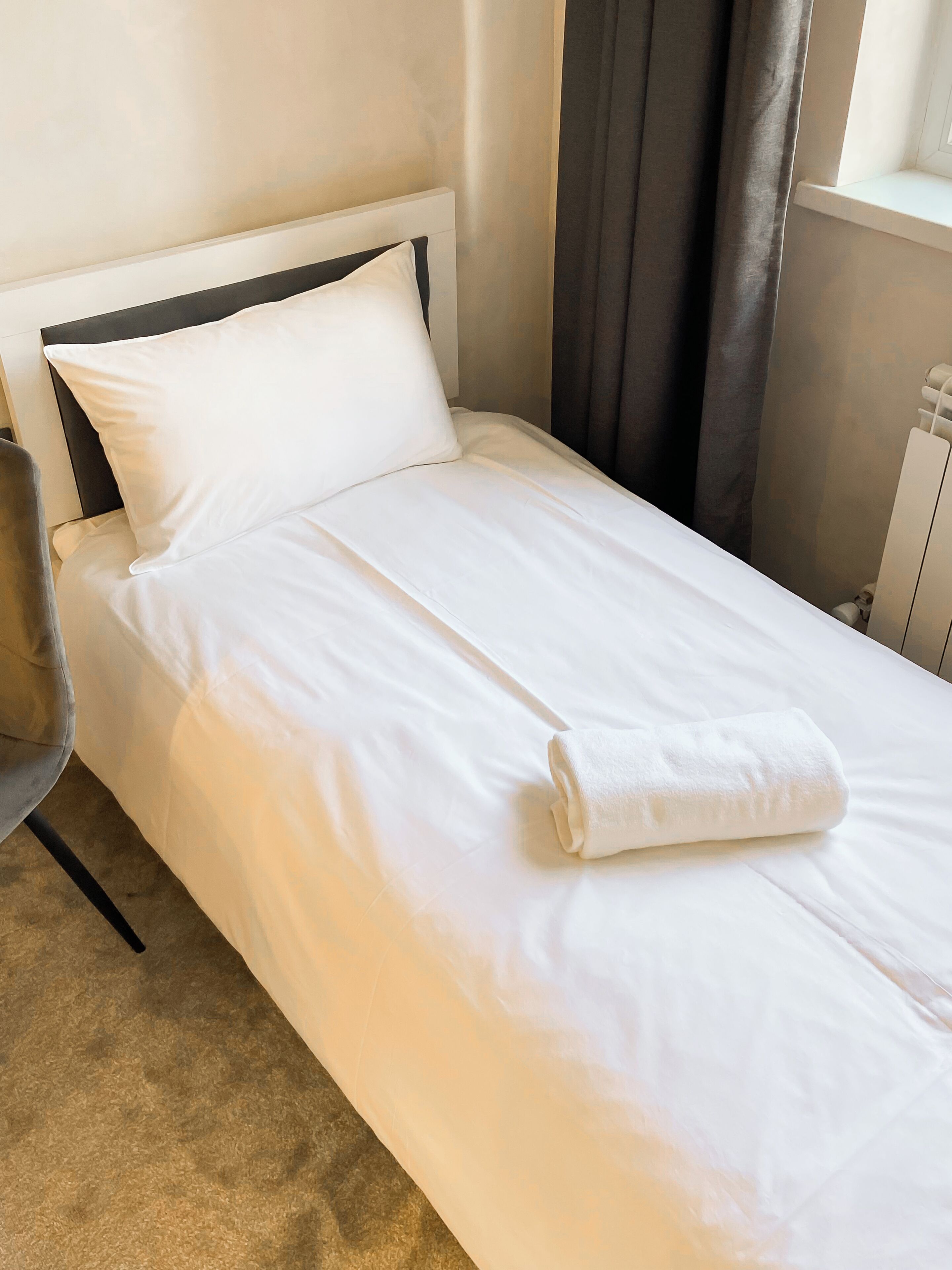 Una cama bien hecha con sábanas blancas, cabecera y una toalla enrollada.