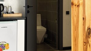 A compact kitchen with a refrigerator, a coffee machine, and a tray with bottles and cups. A door leads to a bathroom with a toilet and a tiled wall. - Beles Apartments (Almaty)