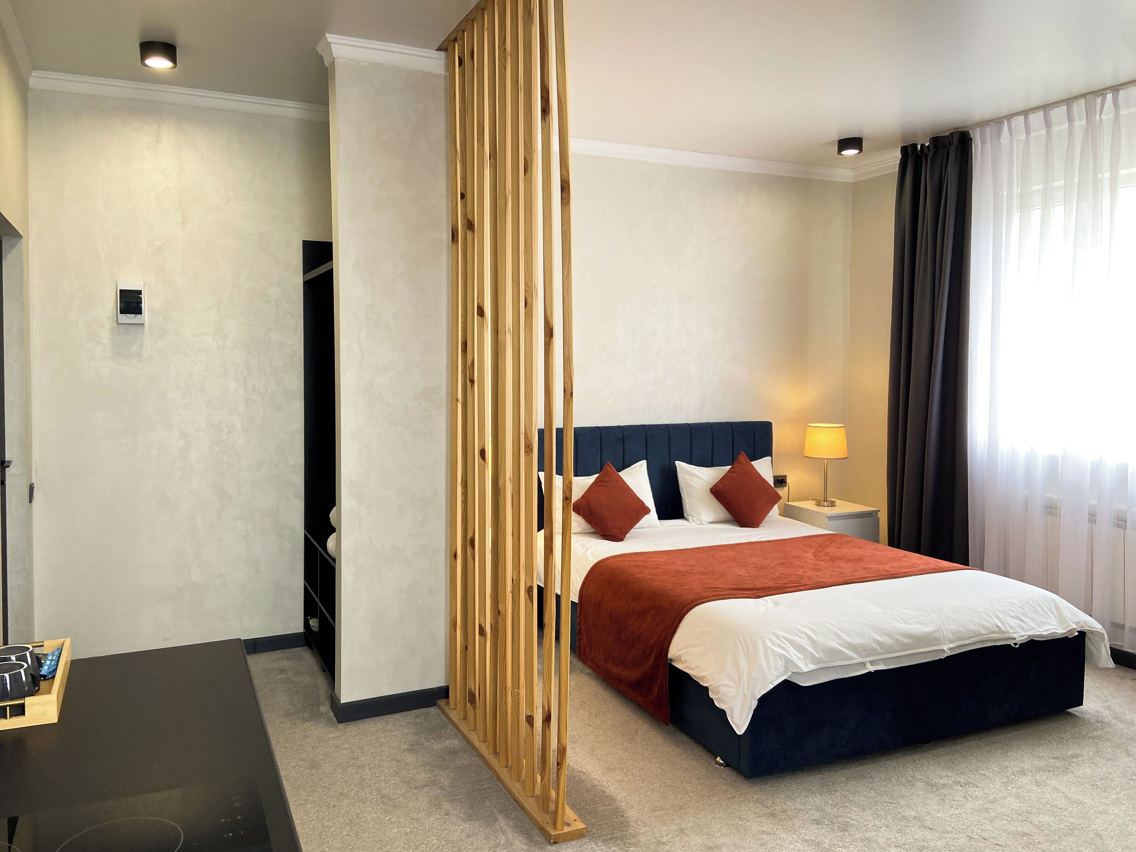 Une chambre d’hôtel avec un lit, une cloison en bois, une table de chevet avec une lampe, et une vue sur l’extérieur par une fenêtre avec des rideaux.