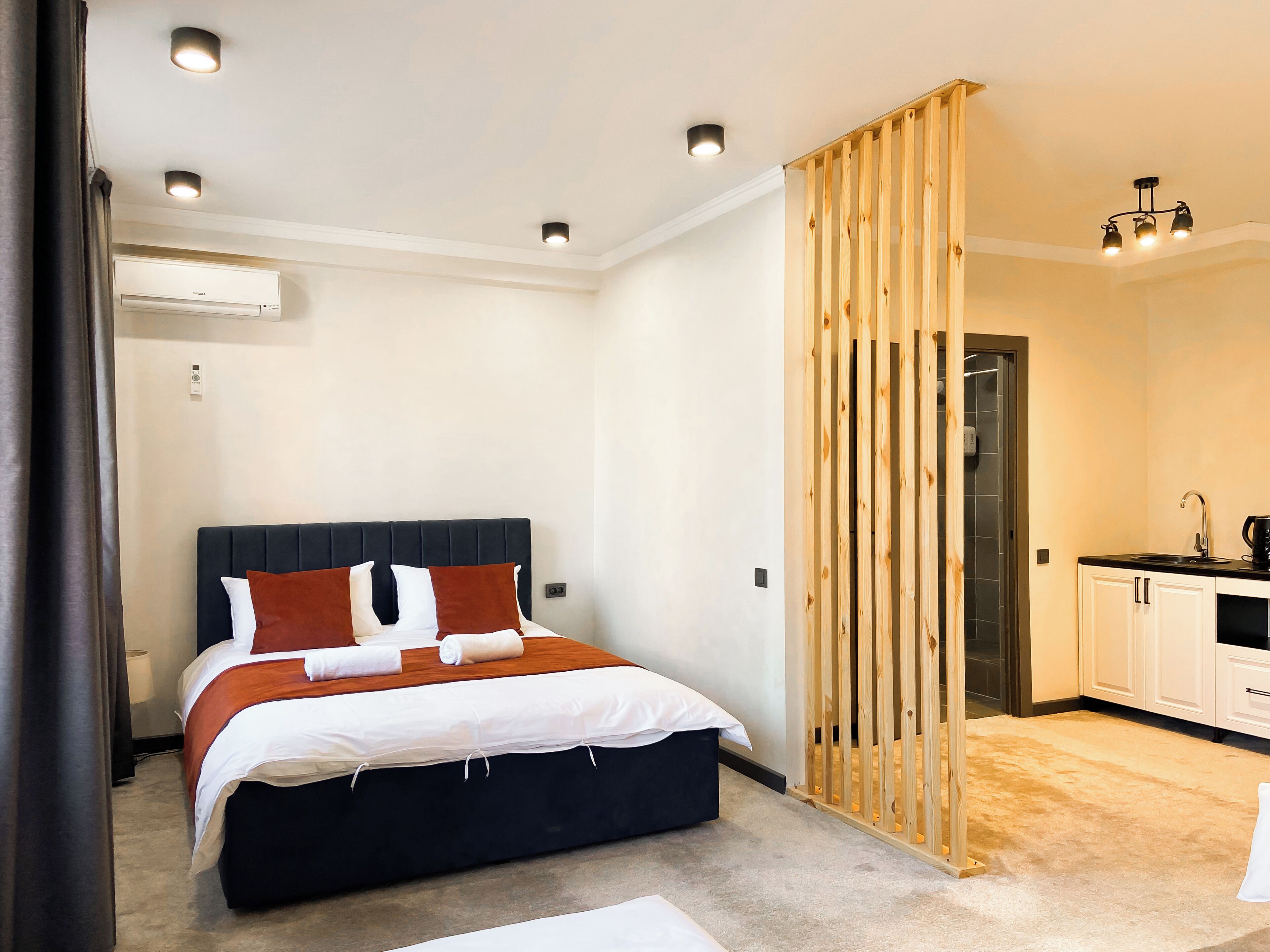 A modern hotel room with a bed, a wooden partition, a kitchen area, and a ceiling light fixture.