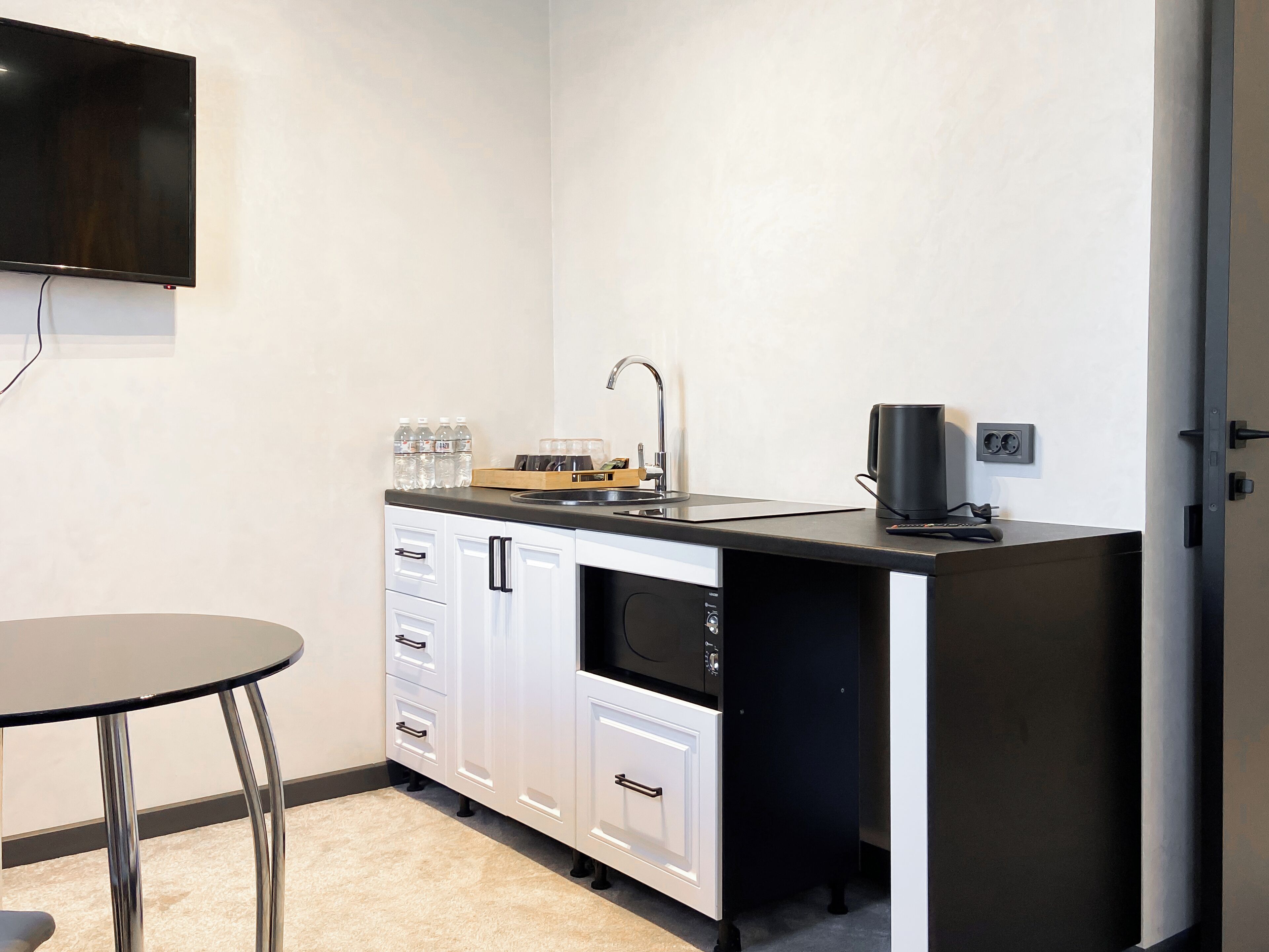 Una cocina compacta con cubierta negra, armarios blancos, un lavamanos y un televisor de pantalla plana montado en la pared.