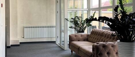 Ein geräumiges Zimmer mit großem Fenster, Heizkörper, einer gesteppten Couch und einer Topfpflanze.