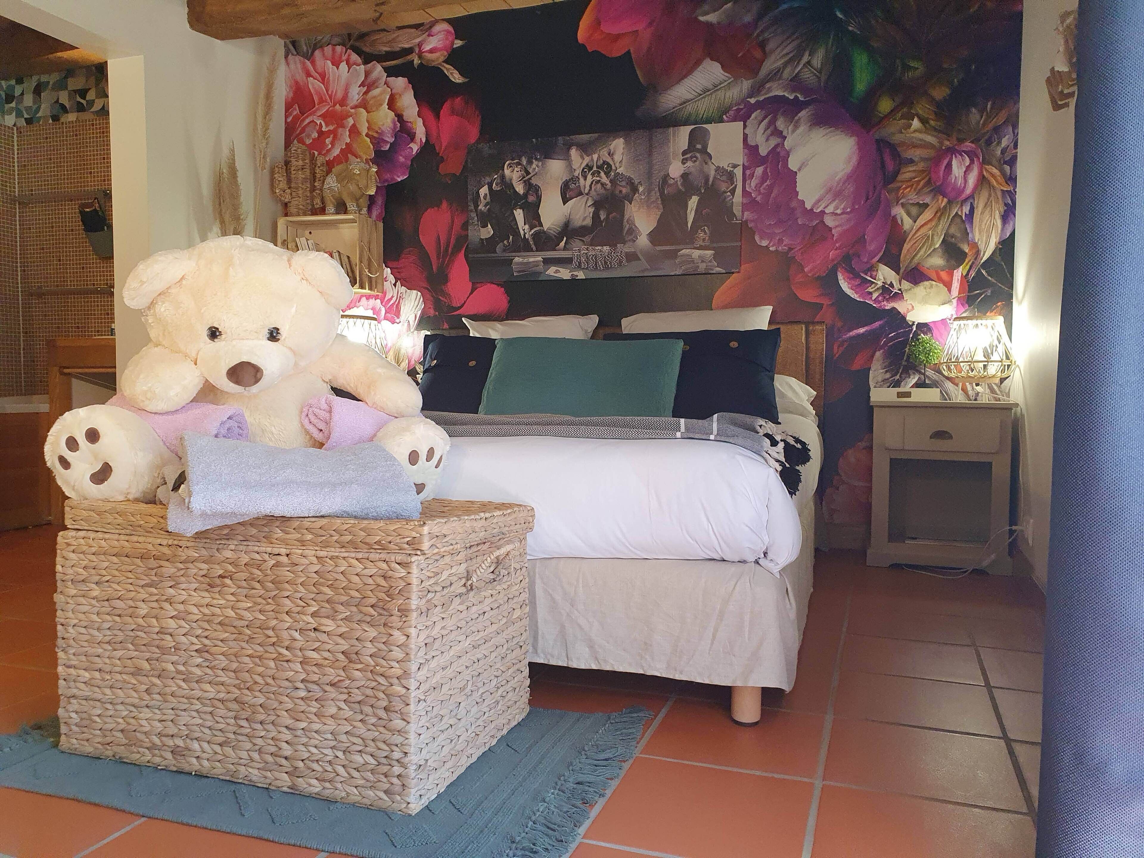 A bedroom with a bed, a large teddy bear on a woven stool, a nightstand with a lamp, and a floral mural on the wall.