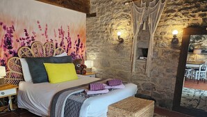A bedroom with a stone wall, a bed with a headboard, bedside lamps, a bedside table, and a view of a dining area. - La Grange Padirac (Padirac)