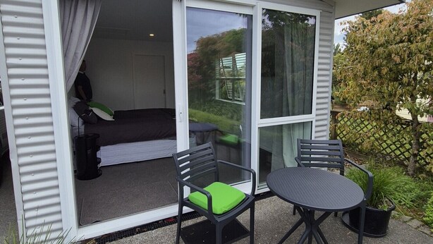 A modern outdoor patio with a sliding glass door leading to a bedroom, two black chairs with green cushions, and a round table. - Private Studio - Perfect for short stays in Taupo. (Taupo)