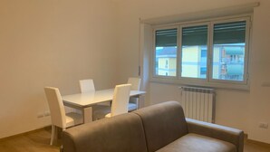 Een kamer met een beige bank, een witte eettafel met vier stoelen en een raam met zonwering.