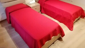 Twee enkele bedden met rode beddengoed, een nachtkastje met een lamp, en een houten hoofdeinde.