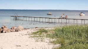En sandstrand med en brygga som sträcker sig ut i stilla vatten, båtar och människor.