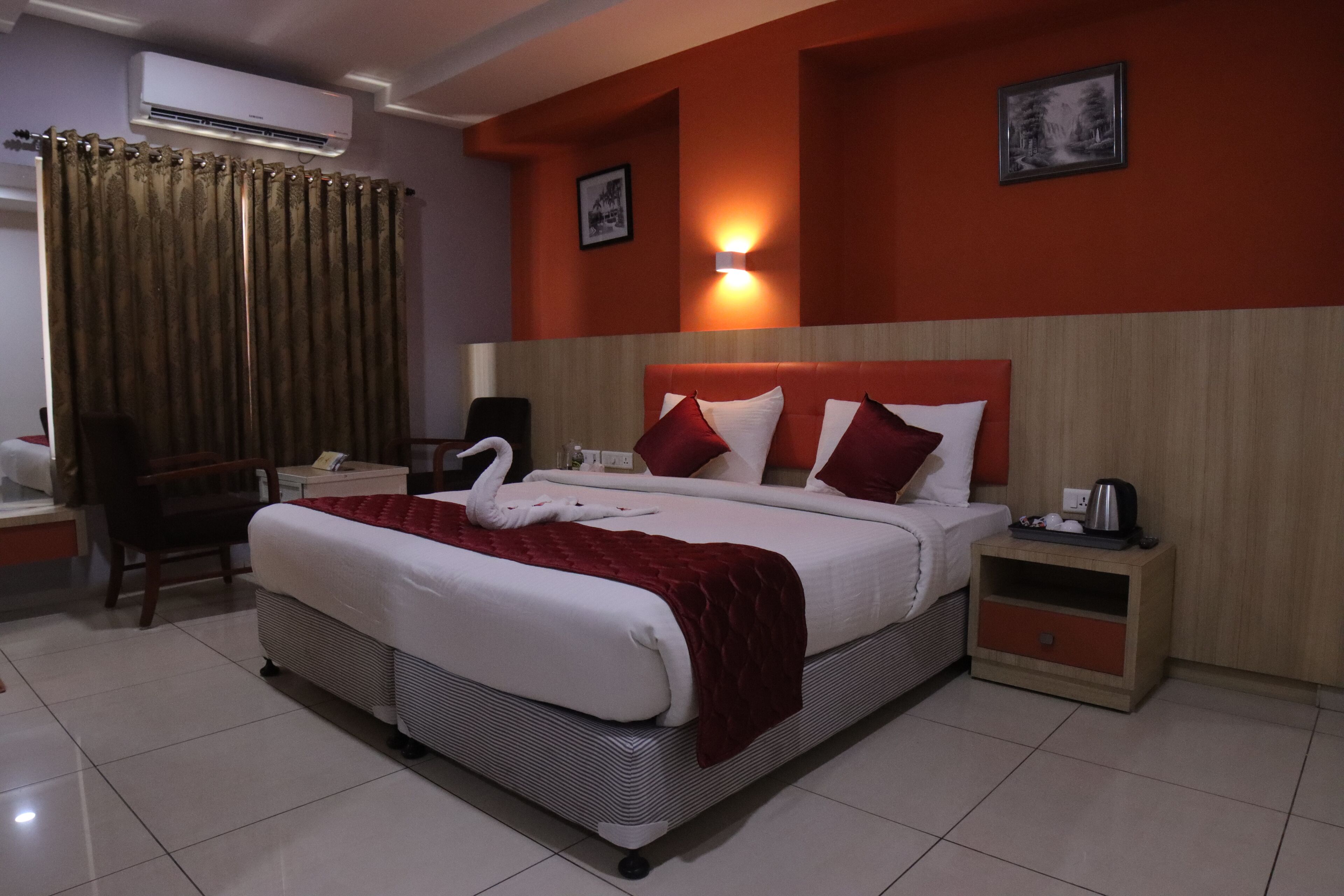A hotel room with a large bed, a desk, a chair, an air conditioner, and a nightstand with a kettle.
